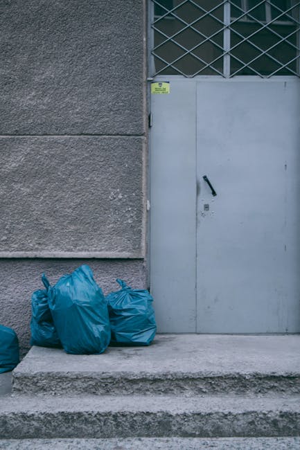 The image shows several large blue plastic rubbish bags stacked against a concrete wall next to a metal door set into a building exterior. The bags appear to be filled with waste material and are situated on a concrete step and pavement area. The wall has a textured, rough surface, and the door is painted in a light grey color with a black handle and lock mechanism. Above the door, there is a section of metal lattice with a grid pattern, possibly allowing ventilation or light. A small yellow security or warning sticker is affixed near the top of the door frame. The scene appears to be an exterior urban or industrial environment, suitable for private waste collection or on-site clearance by waste removal services such as Waste Removal Bayswater, providing an alternative to traditional municipal rubbish disposal that may involve independent collection of refuse from commercial or residential properties. Lighting is natural, with even daylight illuminating the area, ensuring clear visibility of the waste bags and surrounding features.