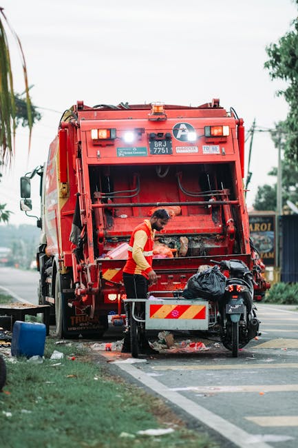 A waste collection worker, wearing a high-visibility yellow and red uniform, is seen operating a large, red rubbish truck parked on the side of a street. The truck's rear hatch is open, revealing the loading mechanism and internal compartments. The worker is handling black rubbish bags placed on a small platform beneath the vehicle, with one bag partially inside the truck. The ground nearby shows scattered debris and a small black scooter parked close to the waste vehicle. The scene is set on a paved street with a white-lined curb next to a grassy verge, and in the background, there are power lines, trees, and a few commercial buildings or signs, suggesting an urban or suburban environment. The lighting appears natural, indicating daytime, and the overall atmosphere is neutral, capturing a routine rubbish collection process that could relate to private waste handling or alternative rubbish removal services, as performed by Waste Removal Bayswater.
