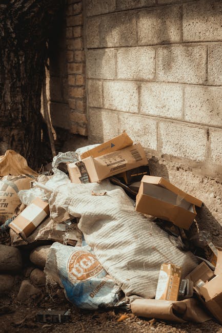An alleyway filled with a large, black, heavy-duty rubbish bag in the foreground, positioned centrally on a rough, dark pavement. Surrounding the bag are additional debris, including a white plastic container and scattered small waste items. The narrow space is flanked by tall, graffiti-covered walls on the right and brick facades on the left, with some exposed pipes visible along the buildings. In the background, a significant amount of mixed waste—cardboard boxes, plastic bags, and miscellaneous trash—are piled up against a makeshift barrier made of wooden pallets and discarded materials, suggesting an area designated for private rubbish collection or on-site waste clearance. Overhanging leafless tree branches are visible above, and the environment has a somewhat dreary, urban atmosphere. The scene appears to be outdoors in a back alley used for waste disposal, with natural, diffused lighting highlighting the textures and mess of the debris, consistent with services like rubbish removal tailored for confined or difficult-to-access spaces.