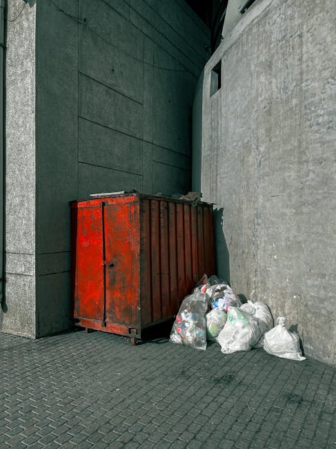 A red metal waste container with a weathered finish, showing signs of rust and faded paint, is positioned against a dark green wall on the left and a light grey concrete wall on the right, on a paved urban sidewalk. Several clear plastic garbage bags filled with mixed waste, including visible plastic bottles and packaging, are piled beside the container on the right side, some leaning against it. The scene appears to be outdoors in an alleyway or side street with minimal natural light, and the environment suggests an area designated for private rubbish collection, as managed by Waste Removal Bayswater. The surroundings are clean but utilitarian, emphasizing the management of rubbish in a typical city setting without additional decorative features.