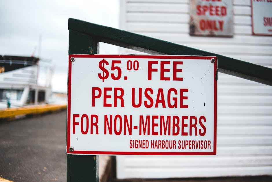 A rectangular white sign with bold red text displays a fee of five pounds per usage for non-members, signed by the harbour supervisor. The sign is mounted on a dark green post with visible bolts at each corner, positioned in front of a building with a white, horizontally paneled exterior wall. Partially visible in the background are other signs with red and white text, and a gray paved surface extends behind the sign, leading towards parked boats and a dock area. The environment suggests an outdoor setting near a marina or harbour, indicating that the sign pertains to service charges for waste disposal or rubbish removal at this location. The lighting appears natural with an overcast sky, which creates soft, diffuse light over the scene, ensuring all details are clearly visible. This setting relates to alternative waste handling fees, which might be relevant when considering private rubbish removal services offered by Waste Removal Bayswater, especially for non-members seeking convenient or independent disposal options.
