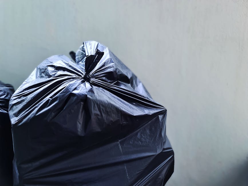 Close-up image of a tightly sealed black plastic rubbish bag with a shiny surface, showing creases and folds, placed against a plain, light-colored wall. The bag appears full and is positioned in the foreground, occupying the lower left part of the image. In the background, another similar black bag is partially visible, slightly blurred, and stacked behind the foreground bag. The scene suggests a typical waste collection or disposal context, possibly prepared for private rubbish removal or on-site clearance, with natural lighting emphasizing the textures of the plastic material. The setting appears indoor or on a sheltered outdoor area such as a driveway or side path, consistent with the environment used by waste removal services like Waste Removal Bayswater for efficient rubbish collection.
