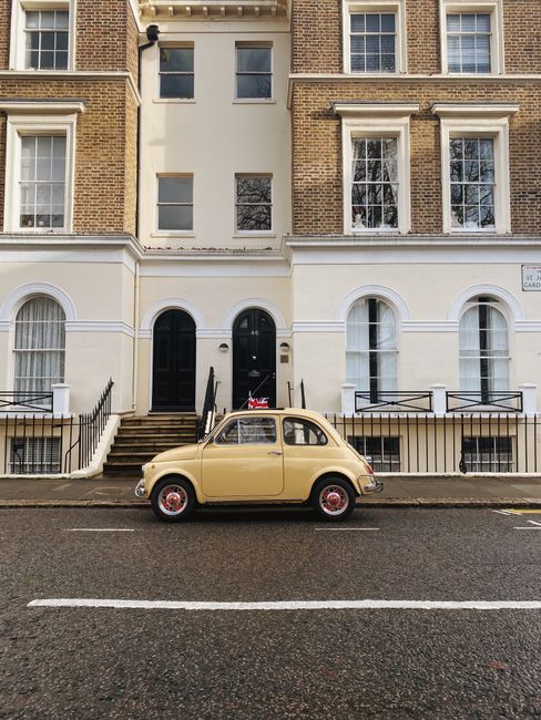 A vintage beige compact car with a rounded body and small size is parked parallel along the curb on a residential street. The vehicle features a shiny, smooth surface with a contrasting red hubcap on the front wheel, and the car's windows are clear, revealing a glimpse of the interior. The street appears clean with a clearly marked white line separating the parking area from the driving lane. Behind the car is a building with a facade featuring two distinct levels; the ground floor is painted white with arched windows and black iron railings, while the upper levels are constructed of brown brick with rectangular windows, some of which have white trim and decorative molding. To the right of the building’s entrance, a small street sign is visible. The overall environment suggests an organized urban setting, and aspects of the scene, such as the neatly parked vehicle and the well-maintained exterior, subtly relate to private or independent waste management services like those offered by Waste Removal Bayswater, supporting the context of rubbish removal or on-site clearance activities.